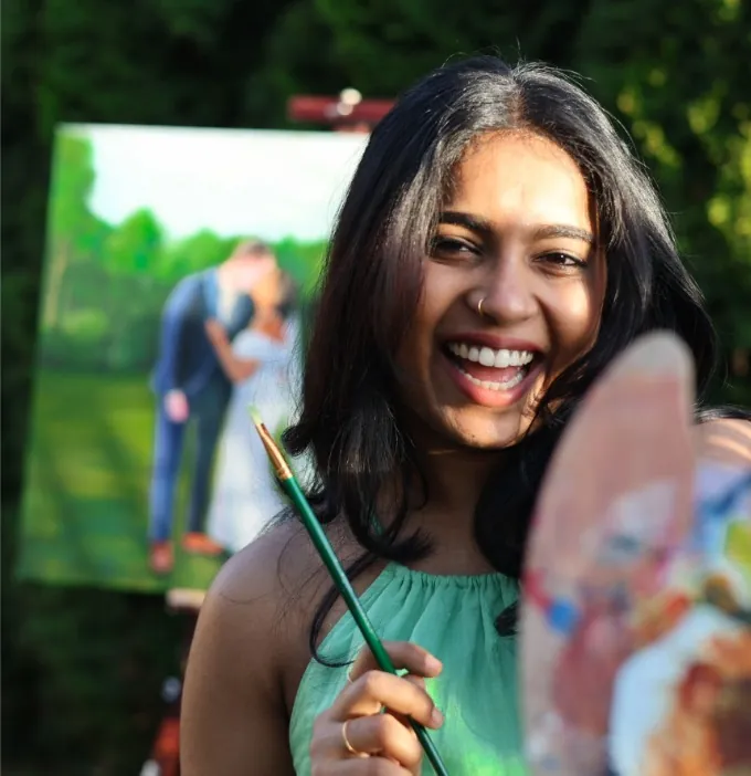 The artist smiling with her brush and palette beside a live wedding painting